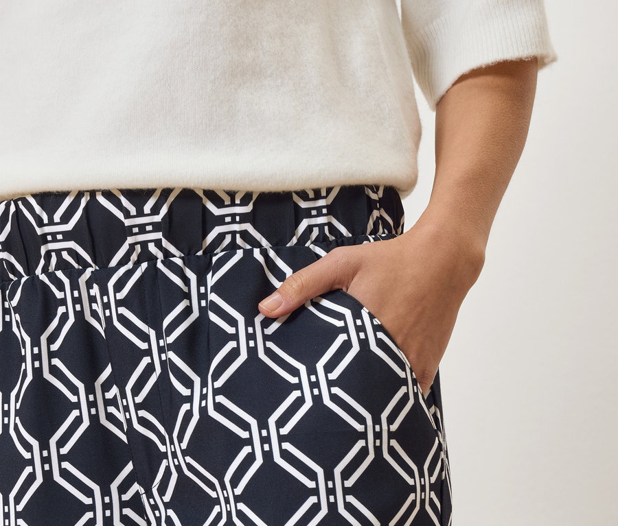 Egy személy közeli felvétele, aki sötétkék, fehér geometriai mintás nadrágot és fehér pulóvert visel, kezével a zsebében.