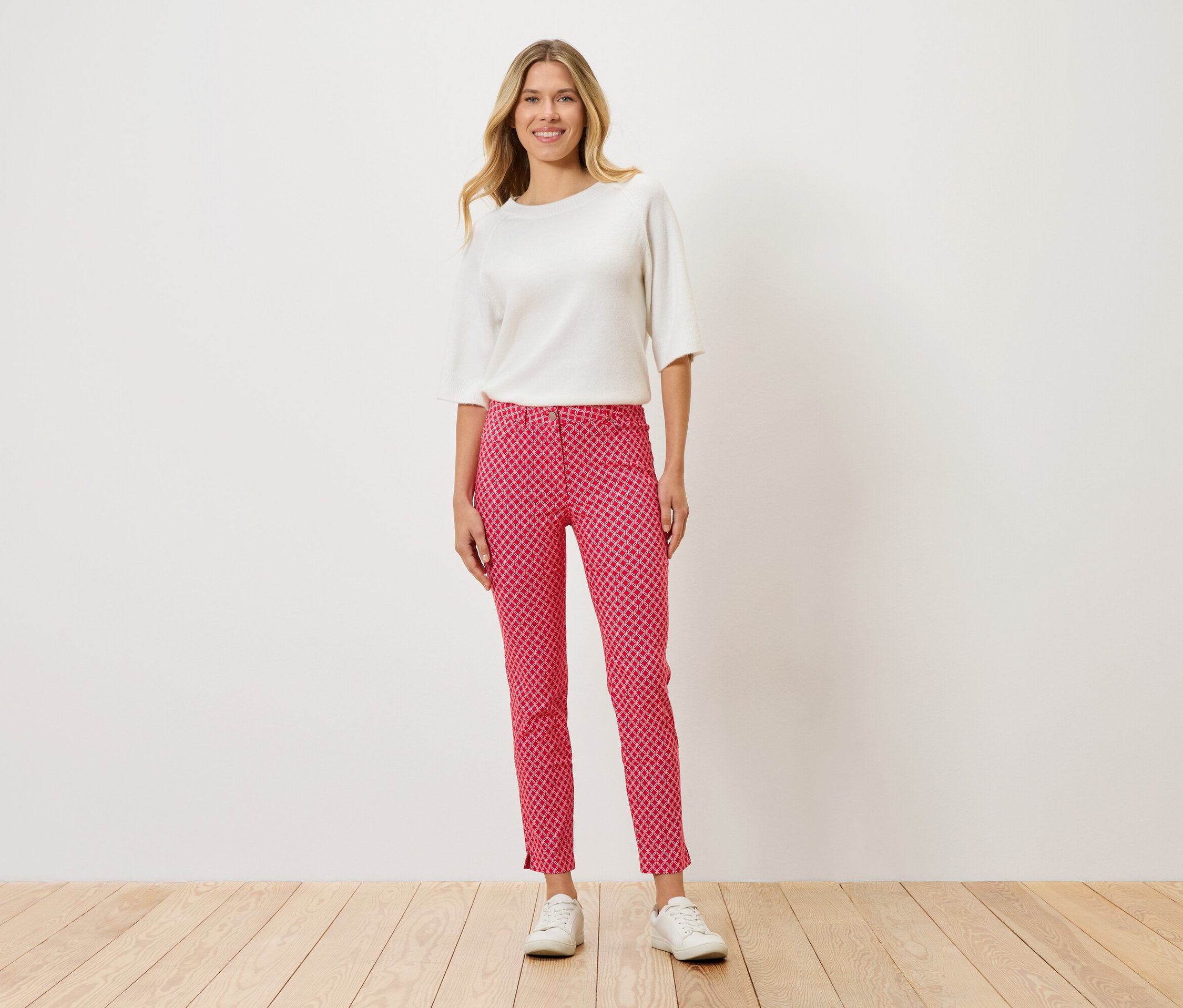 Egy nő áll, fehér felsőt, piros pöttyös nadrágot és fehér cipőt visel.