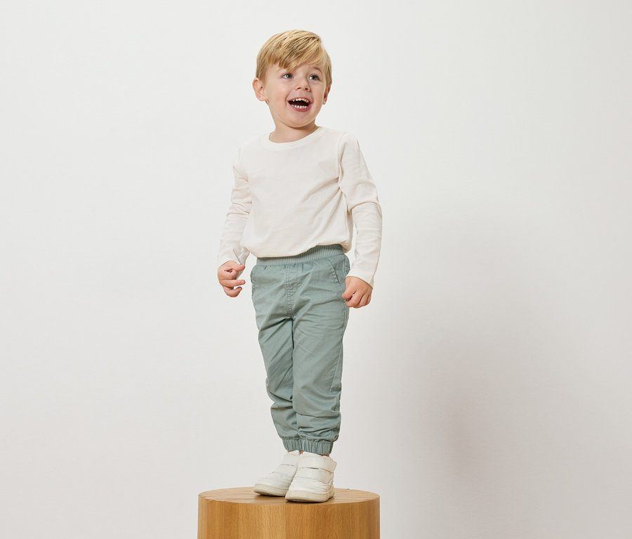 Egy mosolygós, szőke hajú fiú áll egy fa talapzaton, krémszínű, hosszú ujjú felsőt, világoskék nadrágot és fehér tornacipőt visel.