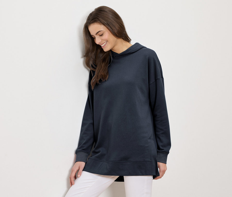 Hosszú, barna hajú nő hosszú, sötétkék kapucnis pulóverben és fehér nadrágban mosolyog, és egy fehér falnak dől.