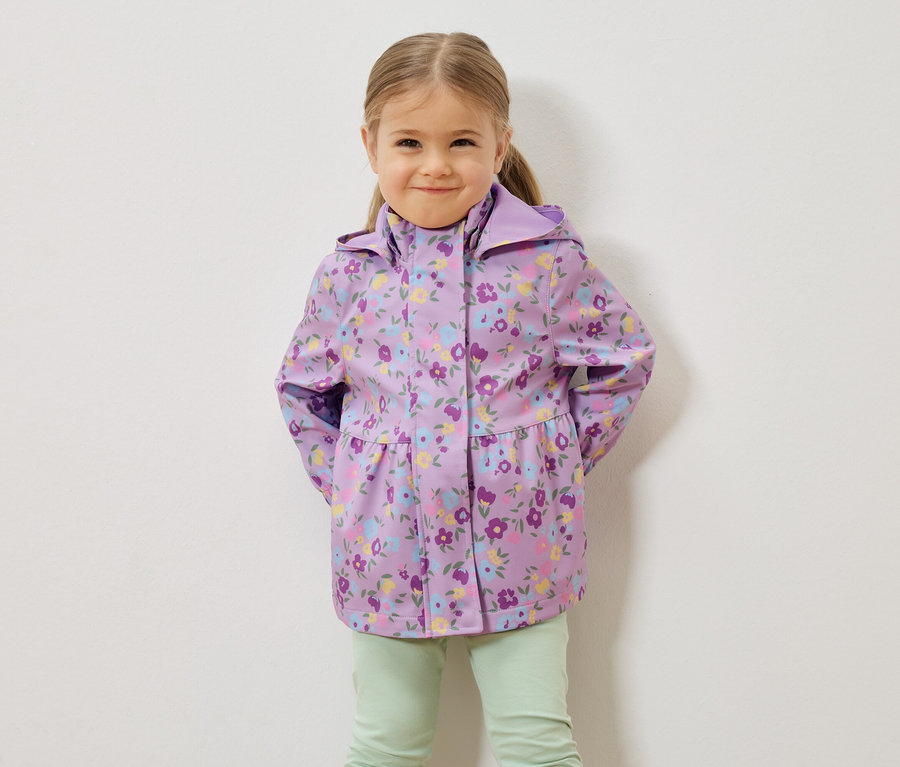 Egy szőke hajú kislány lila, virágmintás esőkabátot és világoszöld nadrágot visel egy fehér fal előtt.