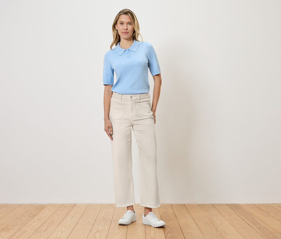 Nő áll a fal előtt, világoskék pólóinget, fehér nadrágot és fehér cipőt visel.