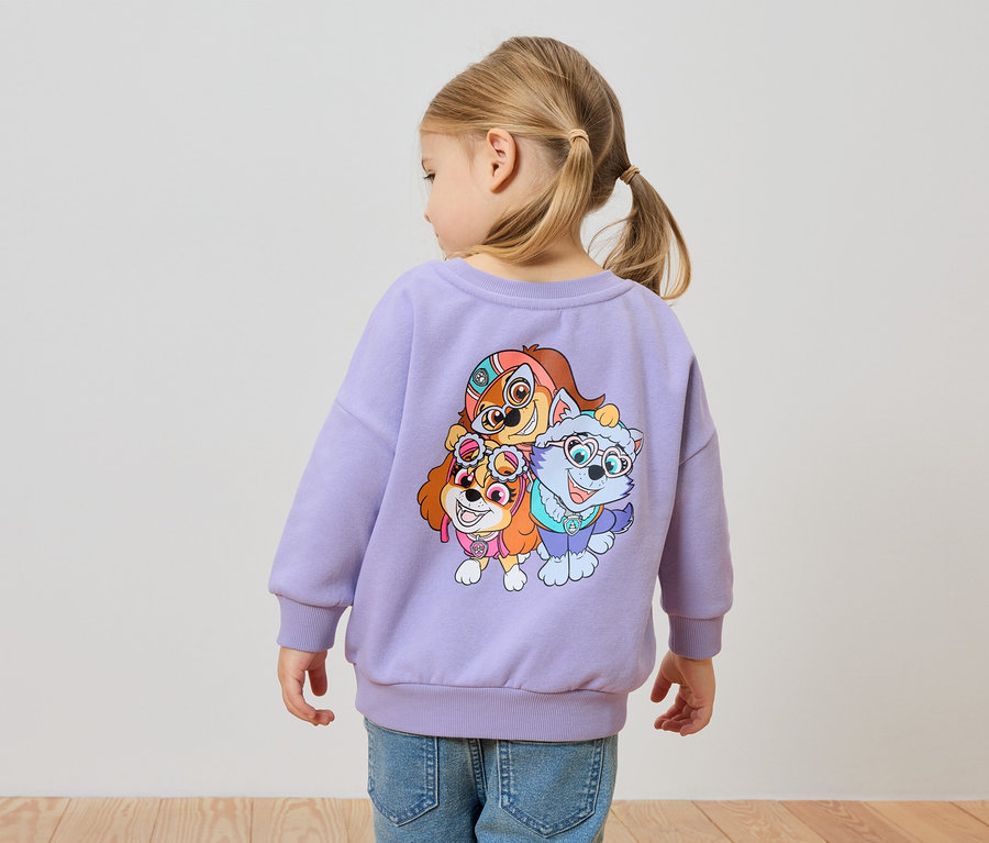 Hátulnézet egy kislányról szőke hajjal, lila Paw Patrol mintás pulóverben és farmerben.
