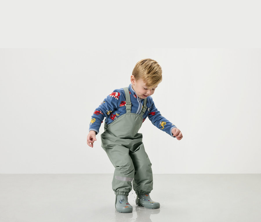 Egy kisfiú zöld esőruhában és gumicsizmában játszik a vízben.