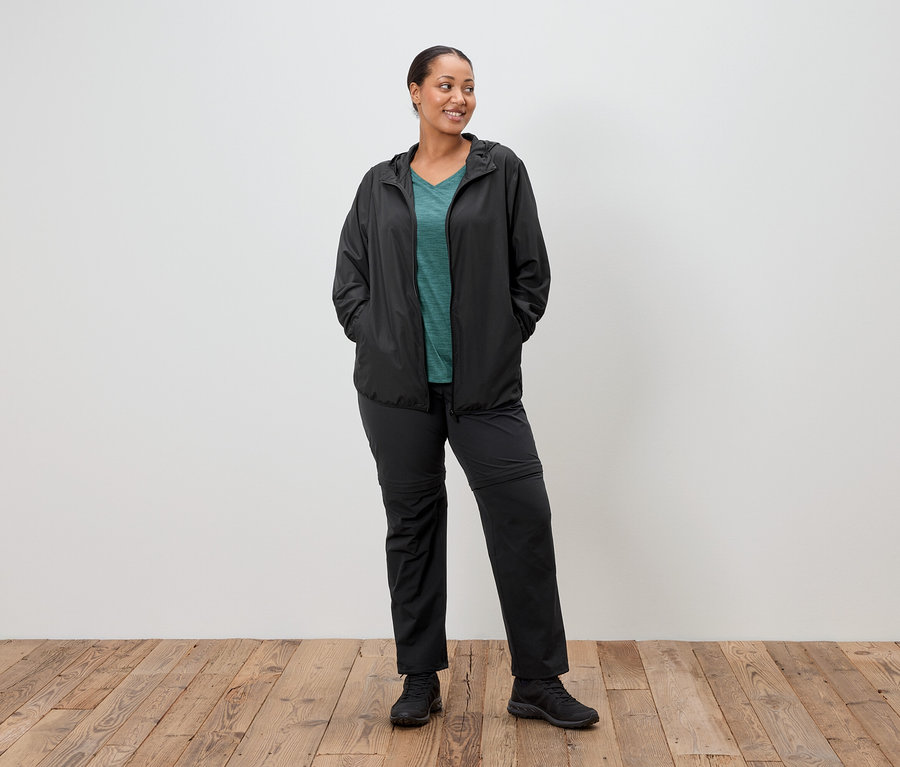 Egy nő áll fekete esőkabátban, zöld felsőben és fekete nadrágban, kezét a zsebében tartva, egy fehér fal előtt.