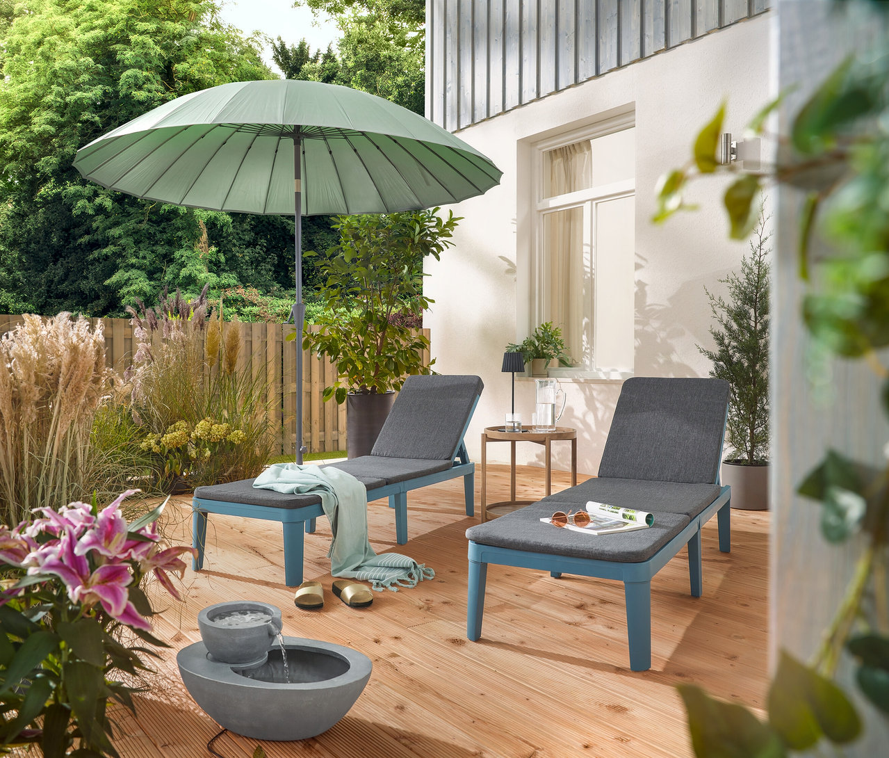 A teraszon két nyugágy áll egy világosszürke Leco Shanghai napernyő alatt és egy napelemes kerti csobogó mellett.