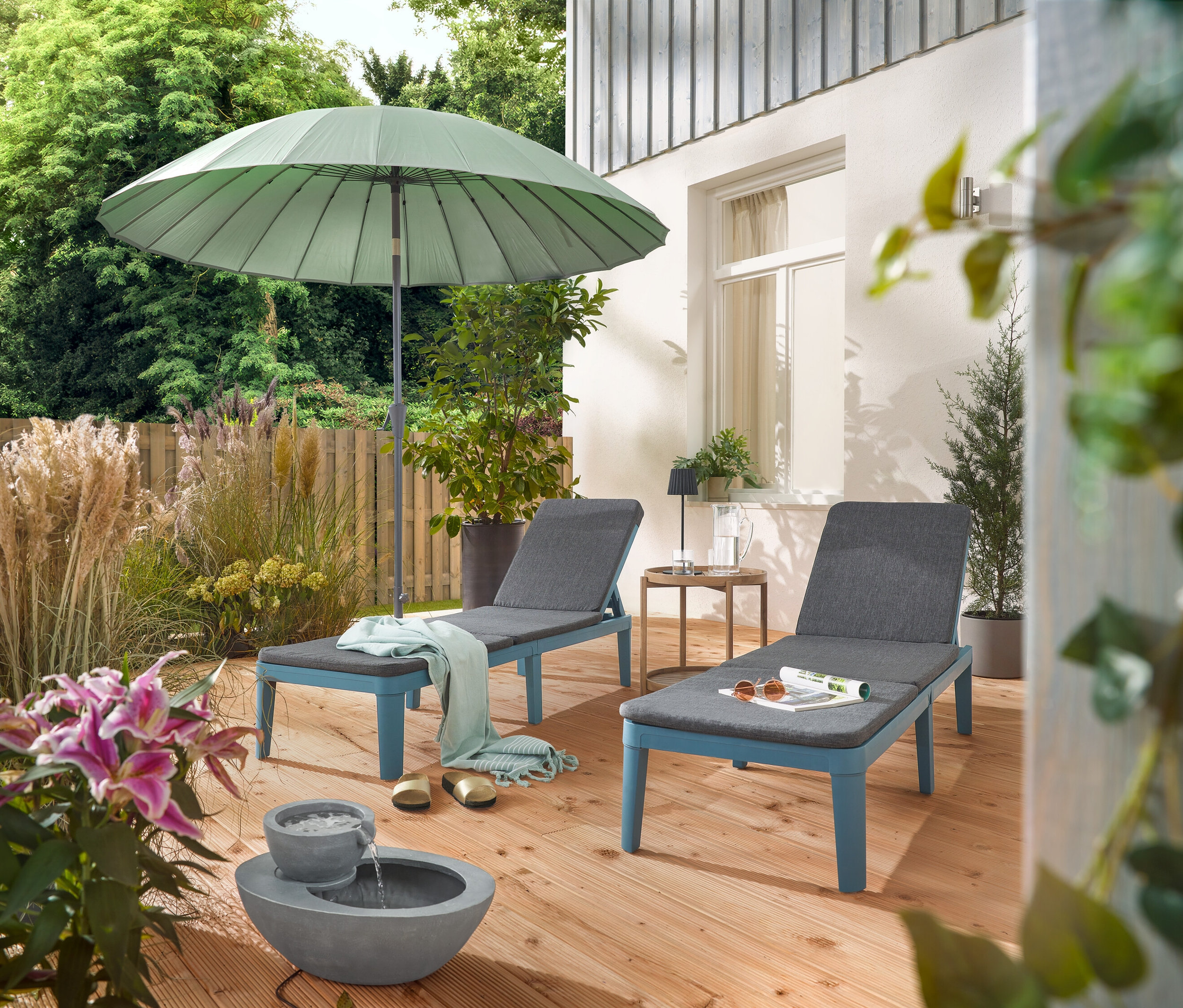 A teraszon két nyugágy áll egy világosszürke Leco Shanghai napernyő alatt és egy napelemes kerti csobogó mellett.