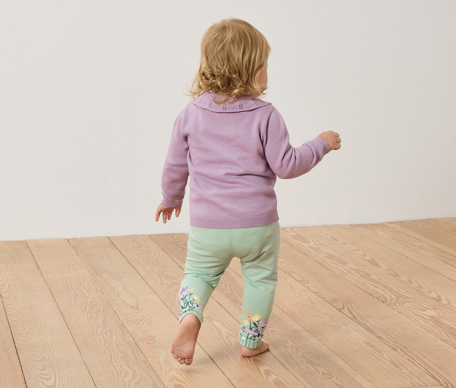 Egy szőke hajú kisgyermek mezítláb áll egy fapadlón, lila pulóvert és világoszöld, virágmintás hímzéssel ellátott nadrágot visel.