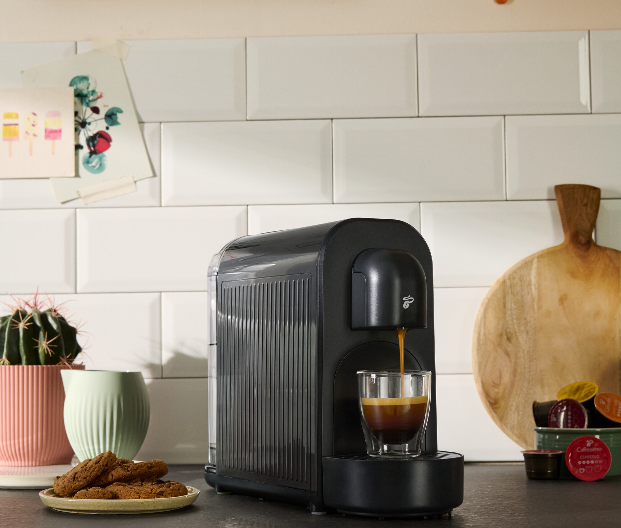 Egy fekete kávéfőző kávéval tölt egy csészét. Kekszek, egy pozsgás növény és konyhai dekorációk is láthatók.