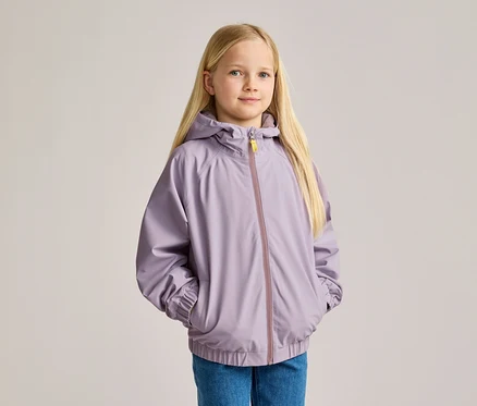 Egy szőke hajú lány farmerben és lila kapucnis kabátban, kezét a zsebében tartja.
