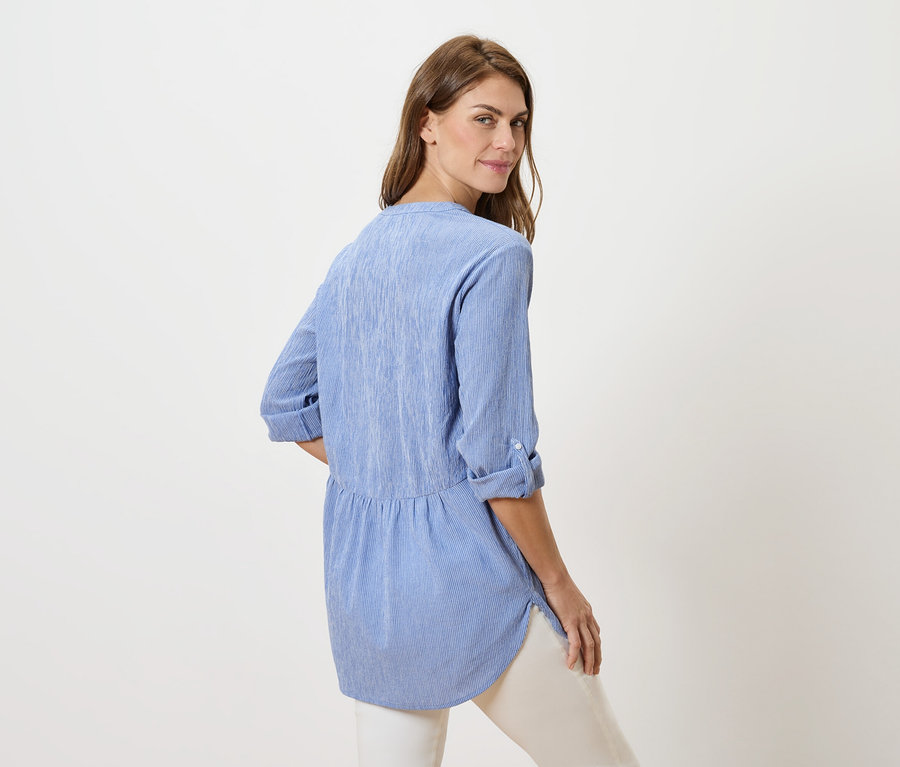 Nő néz a válla fölött, kék blúzt és fehér nadrágot visel.