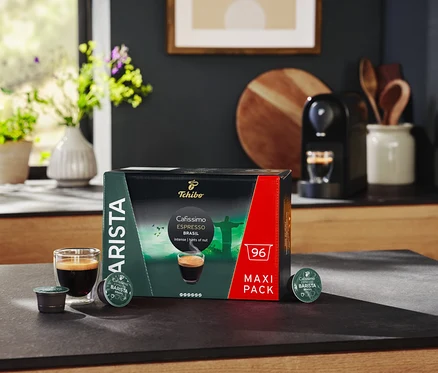 Az asztalon egy csomag Cafissimo BARISTA Espresso Brasil kapszula, egy pohár eszpresszó és egy kávéfőző található.