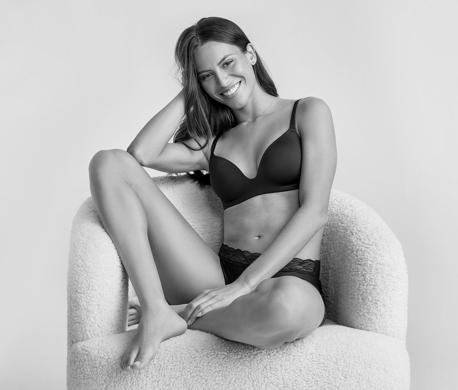 Egy mosolygó, hosszú sötét hajú nő ül egy bolyhos fotelben, fekete puha kosaras melltartót és fekete csipkés bugyit visel. A kamerába néz, egyik karja a fején nyugszik.