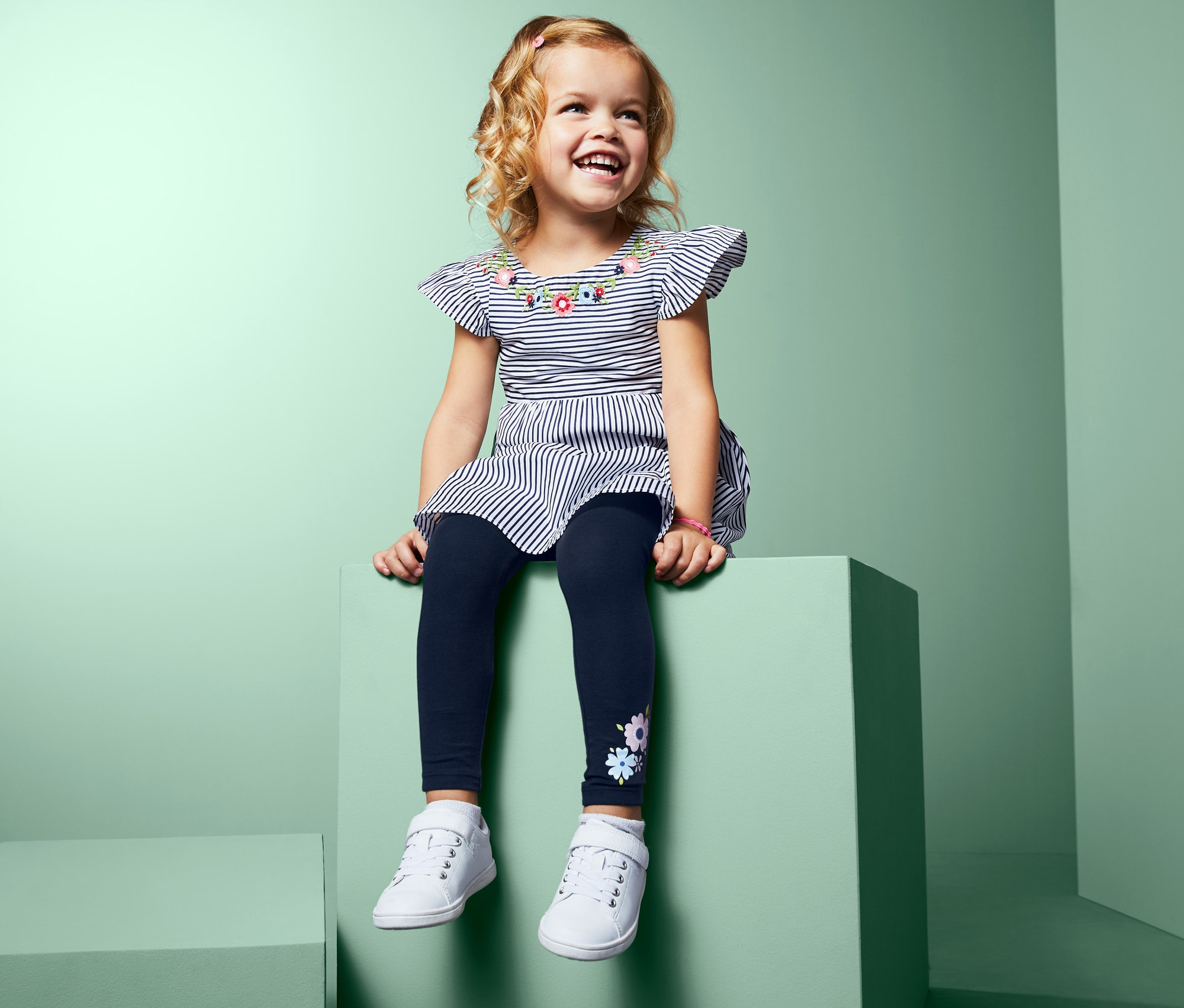 Mosolygó kislány ül egy zöld tömbön, csíkos ruhát és 2 leggings szettben virágokkal.