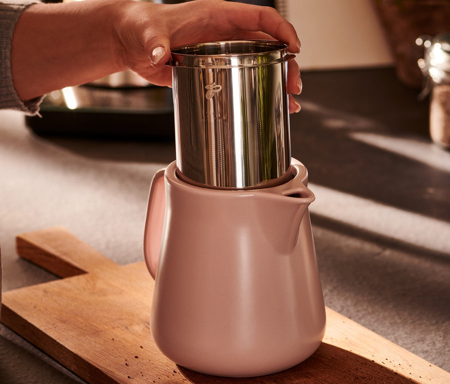Egy kéz egy rozsdamentes acél szűrőt tart egy rózsaszín kerámia kávéskanna fölé.