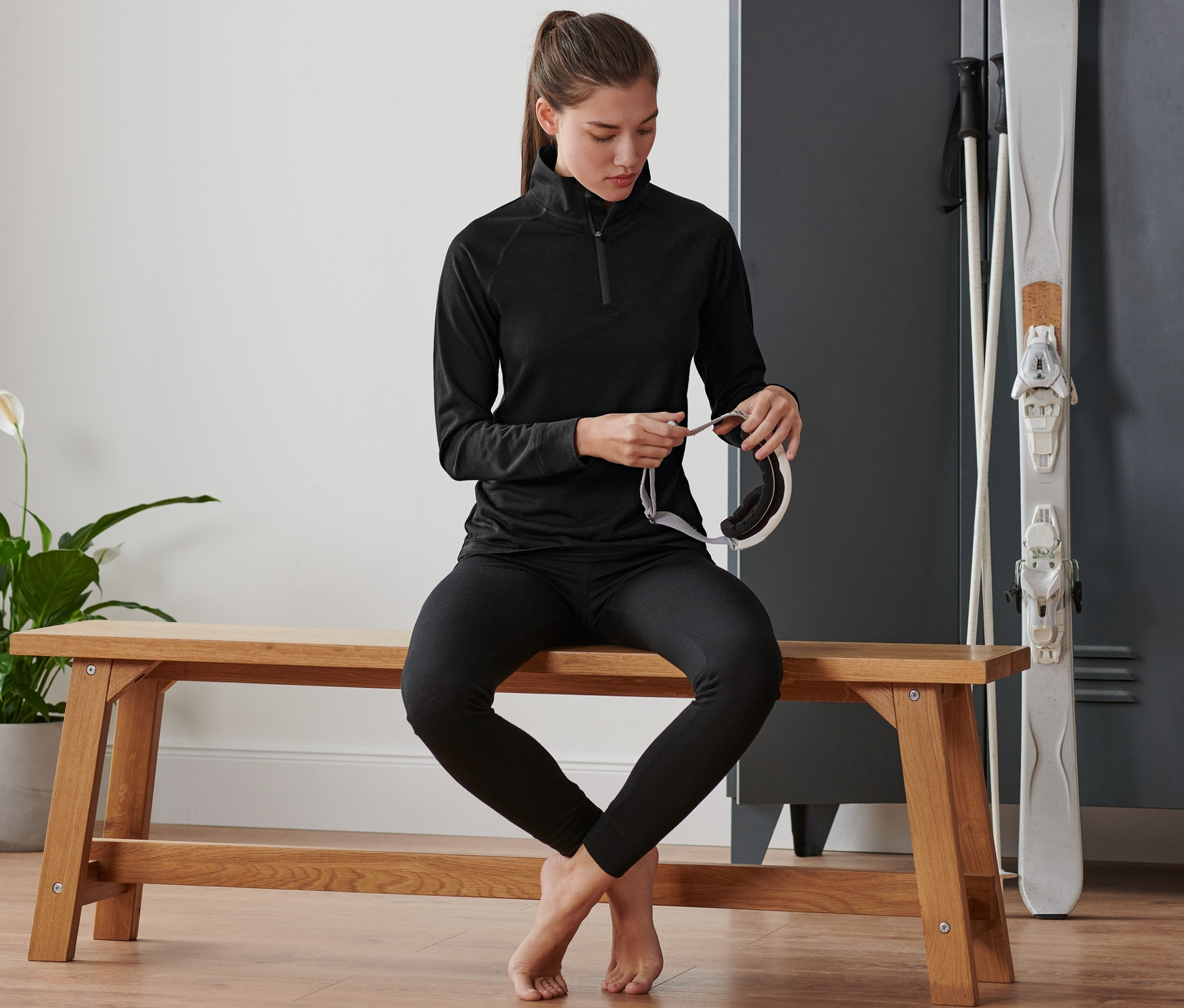 Nő ül egy padon és sí szemüveget tart. Merinógyapjú funkcionális felsőt visel.