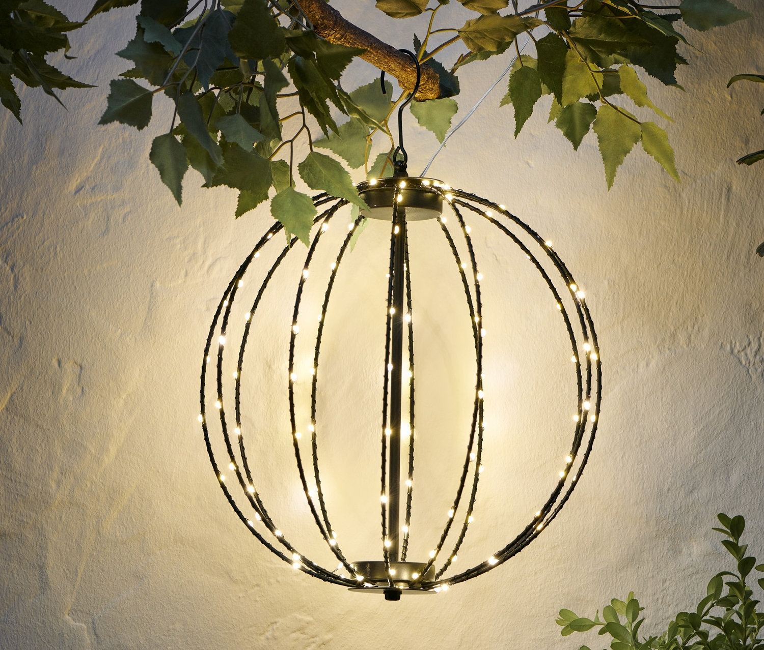 Egy fém LED-es gömb lóg egy ágon egy fehér fal előtt.