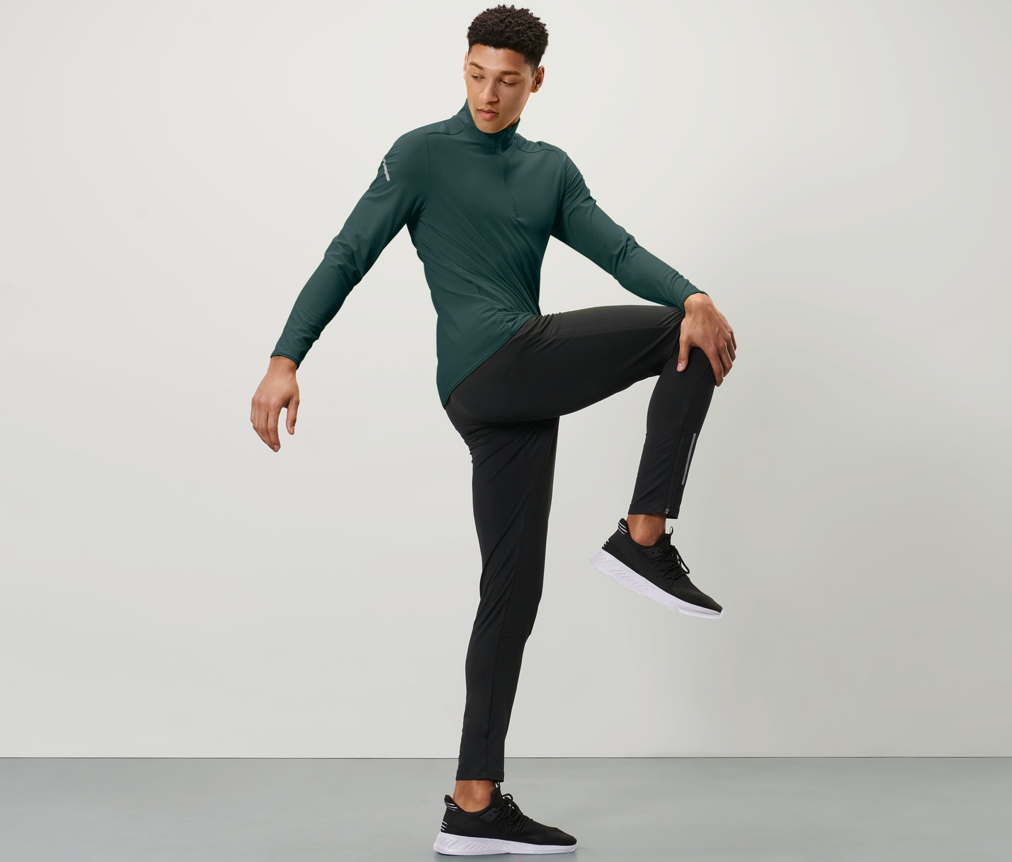 Férfi zöld sportfelsőben és fekete szélálló futónadrágban az egyik kezével a mellkasa előtt tartja a térdét.