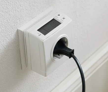 Egy fehér falon egy konnektor található energiafogyasztás-mérővel és egy bedugott fekete zsinórral.