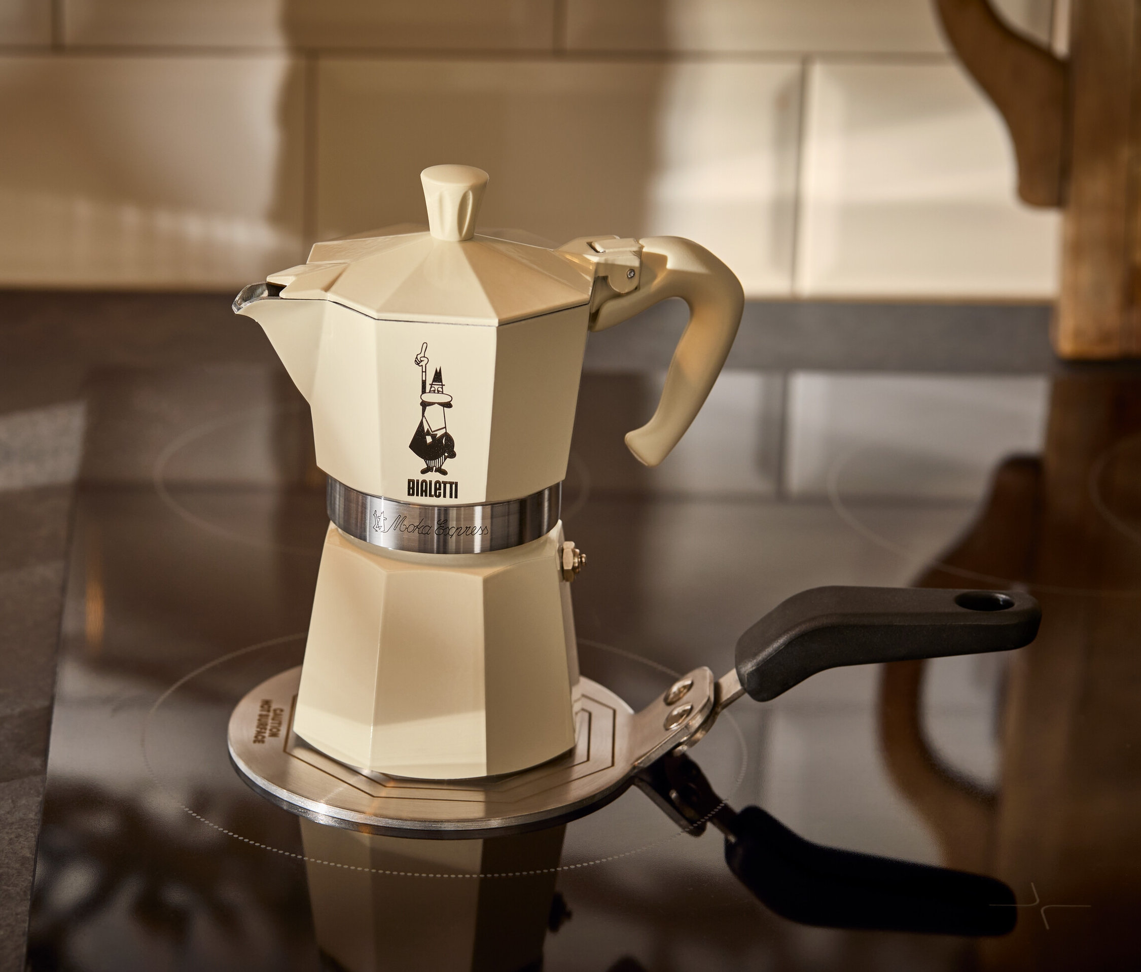 Egy krémszínű Bialetti Moka Express kotyogós kávéfőző 190 ml-es egy Bialetti 13 cm-es indukciós átalakítólapon áll egy presszóspohár mellett.