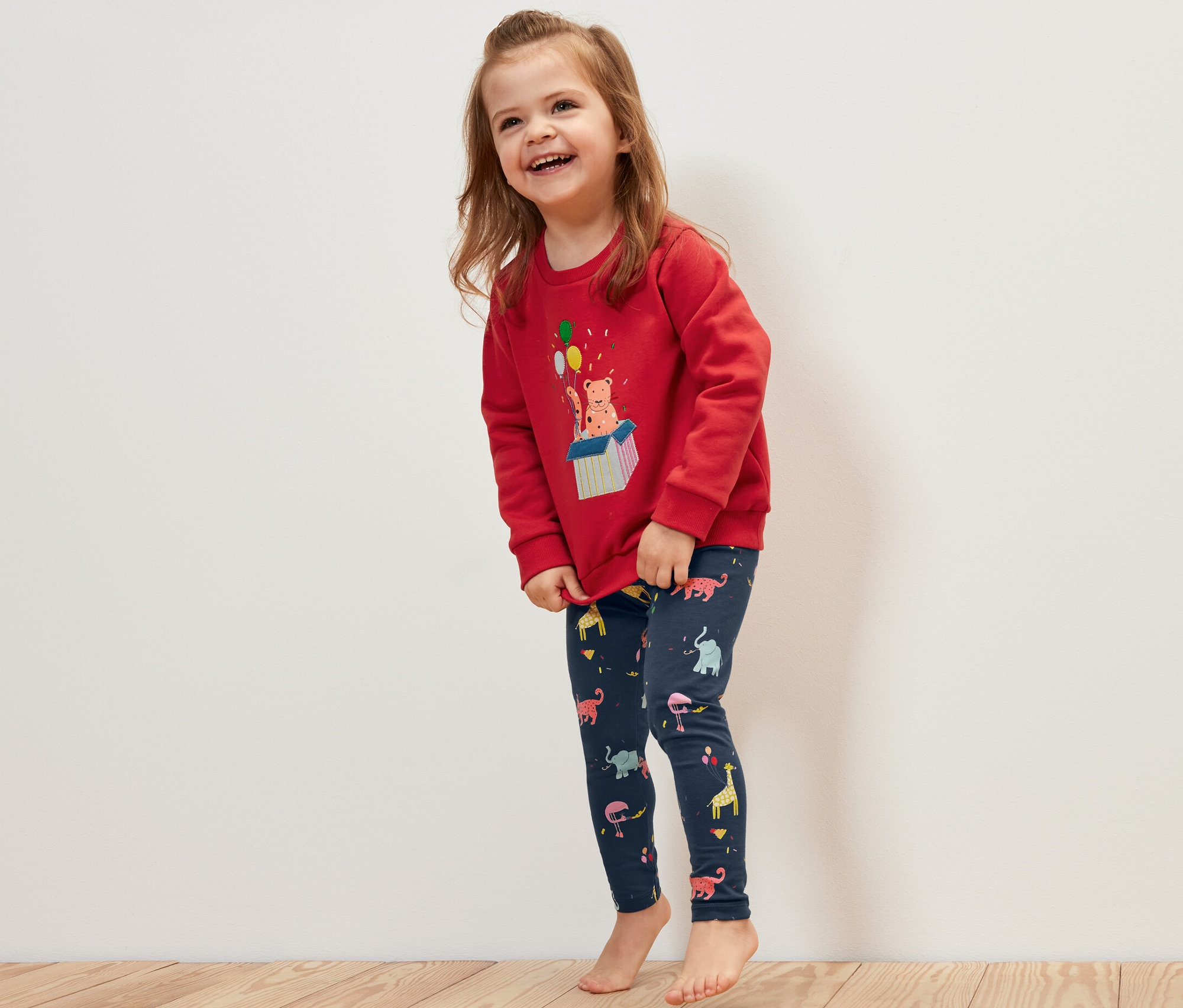 Mosolygó kislány piros pulóvert és állatmintás 5 darabos kisgyerek leggingst visel.