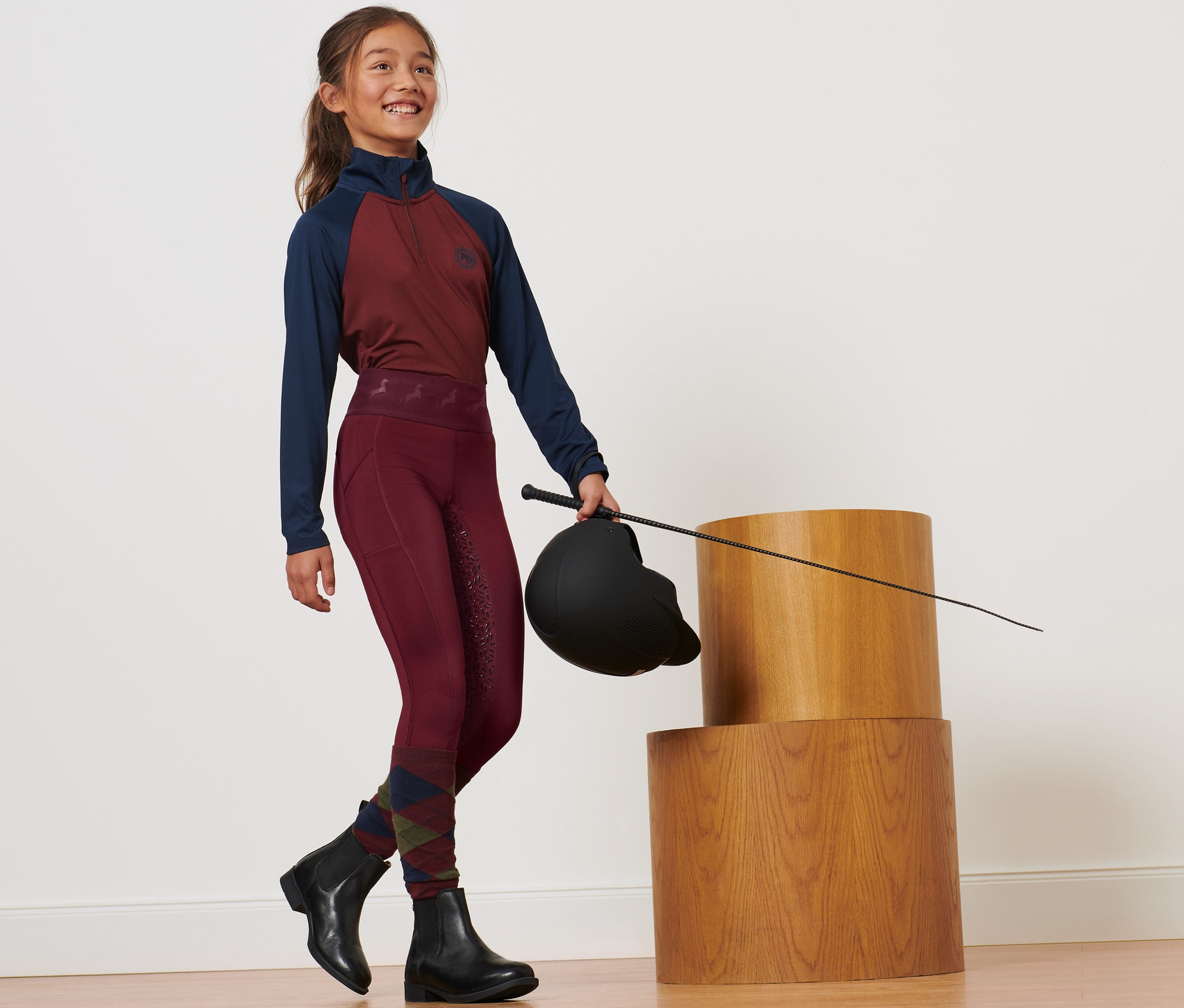 Lány lovaglósisakot és ostort tart, piros, gyerek lovaglónadrágot visel.