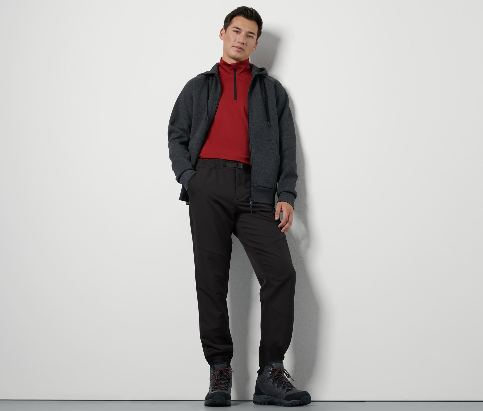 Egy férfi áll és fekete termonadrágot, cipzáras pulóvert és túrabakancsot visel.