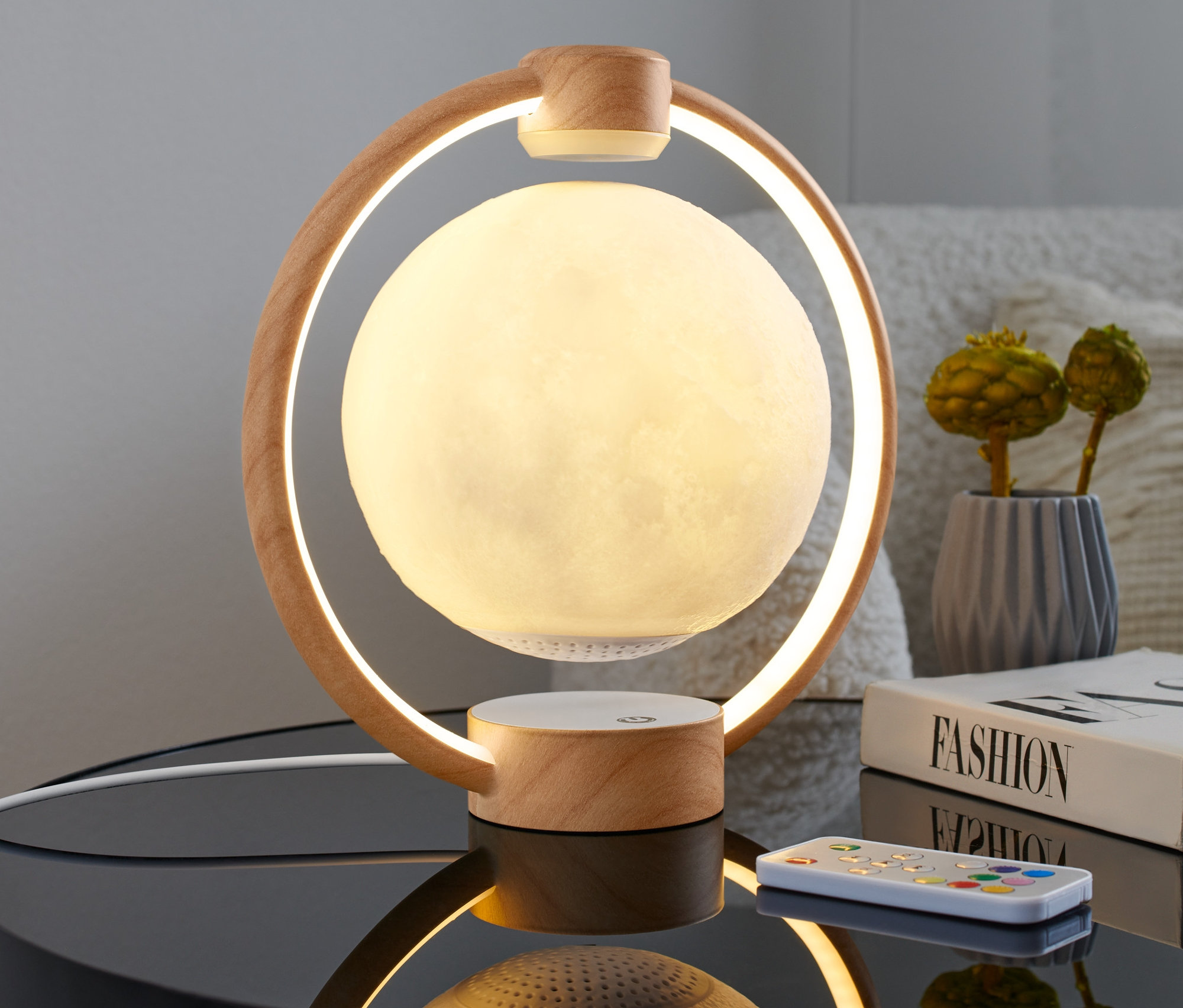 Egy lebegő Hold asztali lámpa, Bluetooth hangszóró egy éjjeliszekrényen áll egy könyv és egy távirányító mellett.