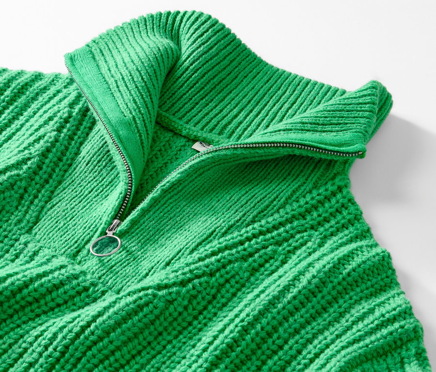 Egy zöld, gyapjú anyagú, állónyakú pulóver részlete, részben nyitott cipzárral.