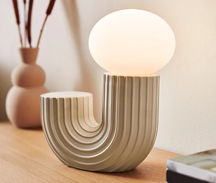 Egy design LED-es asztali lámpa áll egy fa asztalon egy váza és könyvek mellett.