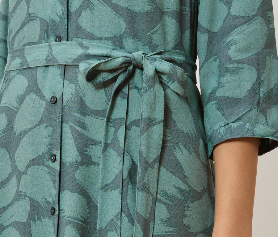 Egy türkizkék, gombos ruha részlete, szürke ecsetvonás mintával és megkötött derékkal.