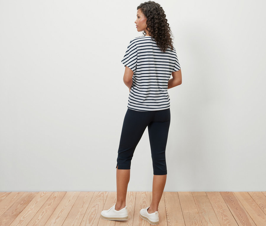 Egy nő háttal áll a kamerának, és egy ejtett vállú, csíkos pólóblúzt, egy sötétkék háromnegyedes nadrágot és fehér tornacipőt visel.