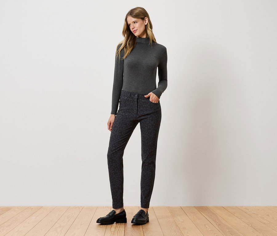 Egy nő áll és pózol szürke garbóban, mintás nadrágban és fekete cipőben.