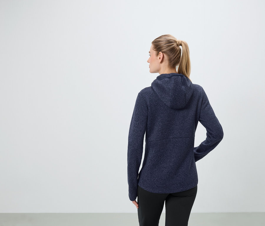 A szőke haját lófarokba fogó nő sötétkék kapucnis pulóvert és fekete nadrágot visel.