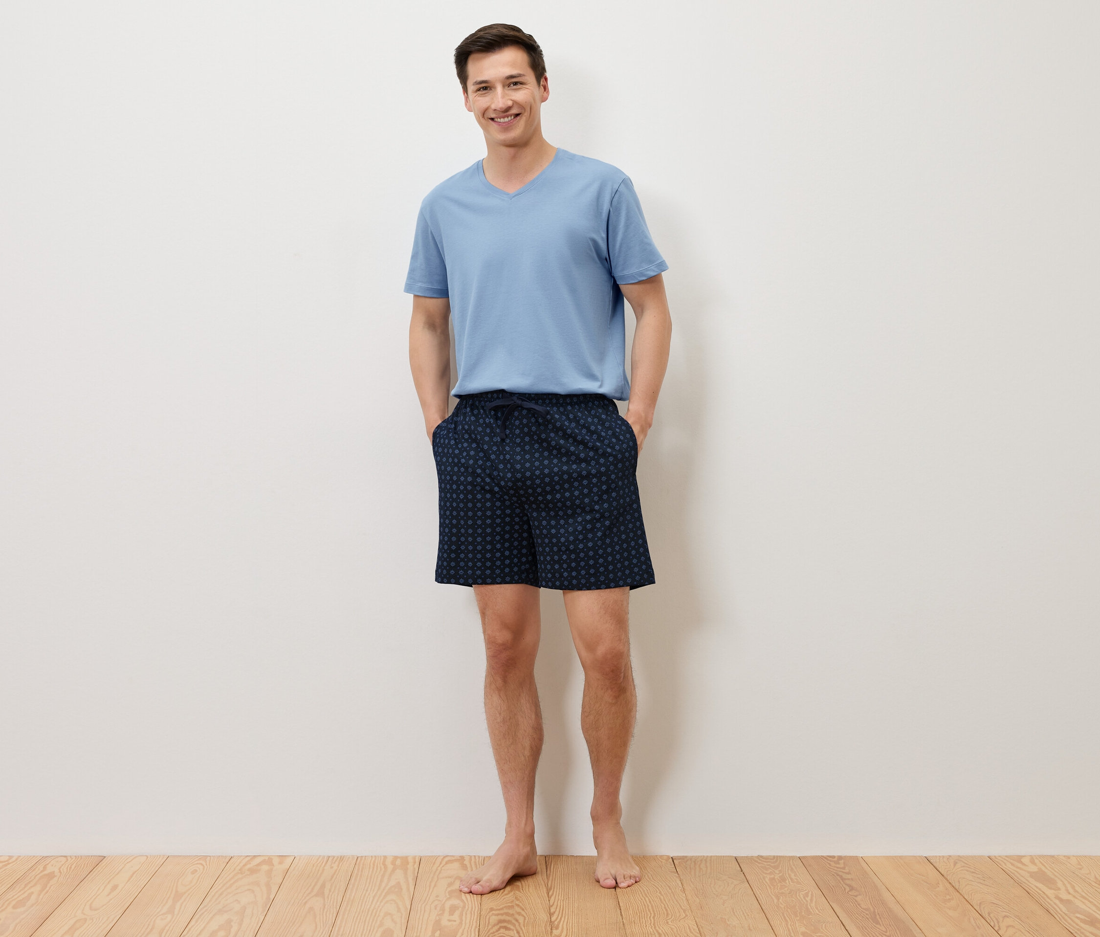 Egy férfi mezítláb áll, kezével a sötétkék pöttyös rövidnadrágja zsebében, és egy világoskék pólót visel egy fehér fal előtt.