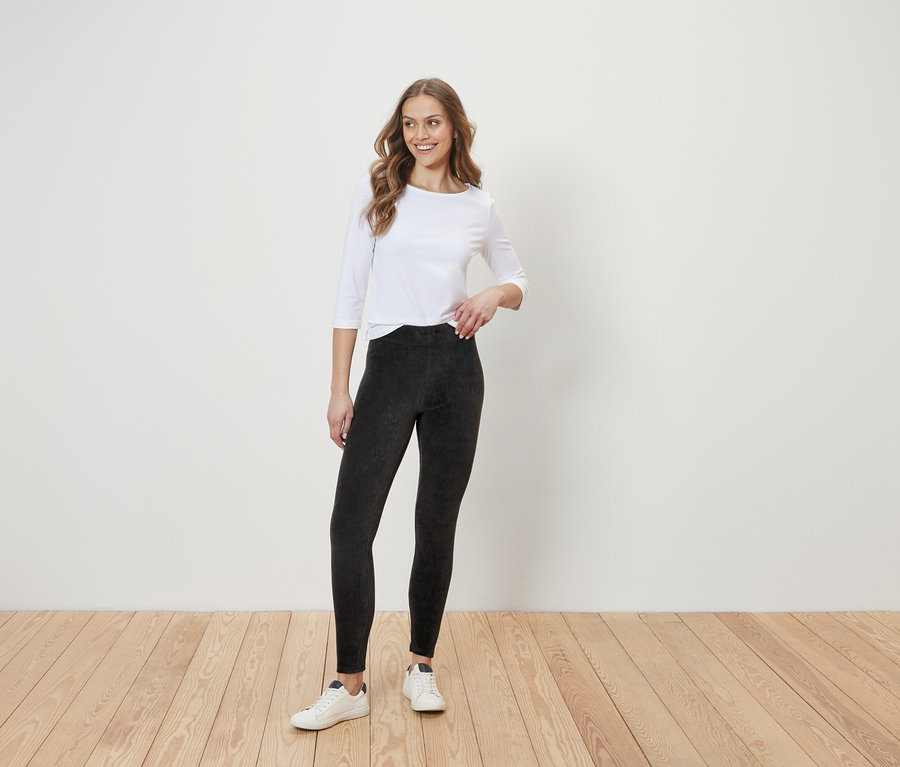 Egy nő áll egy fapadlón egy fehér fal előtt. Fehér, háromnegyedes ujjú blúzt, fekete leggingset és fehér tornacipőt visel.