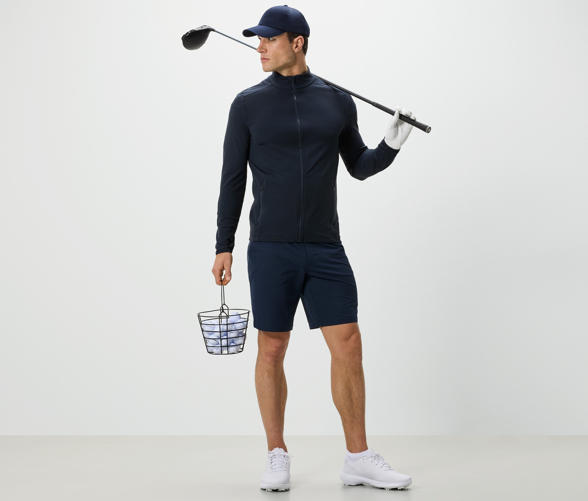 Férfi sötétkék funkcionális felsőben golflabdákkal teli kosarat és golfütőt tart.