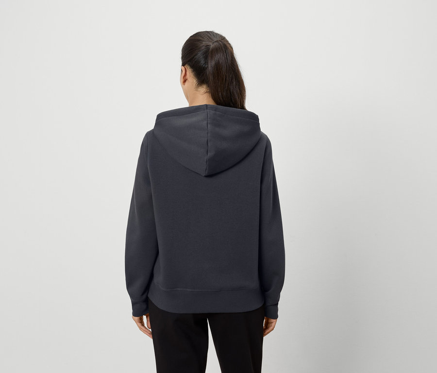 Egy nő háttal áll, sötétszürke kapucnis pulóvert és fekete nadrágot visel.