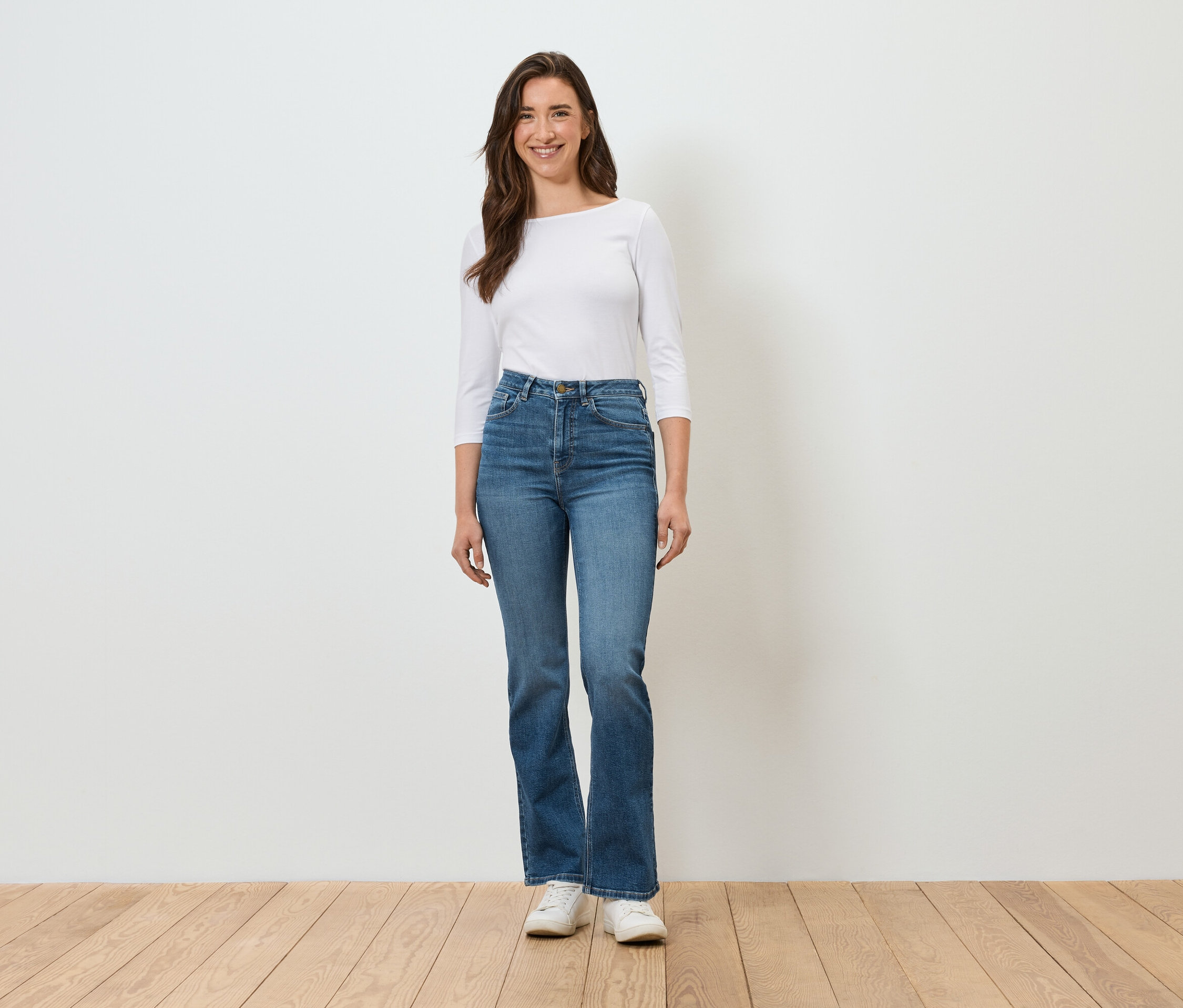 Egy nő áll egy fehér fal előtt, kék farmerben, fehér ingben és fehér tornacipőben.