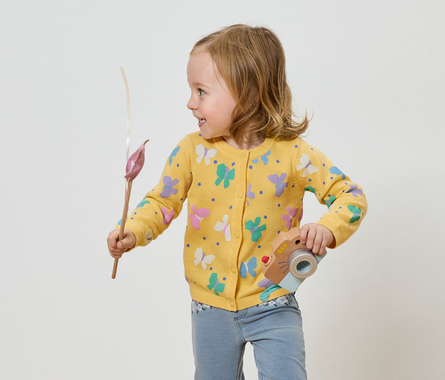 Kislány pillangós sárga kardigánban játék fényképezőgépet és virágot tart.