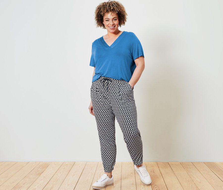 Nő pózol kék V-nyakú felsőben és mintás jersey nadrágban.