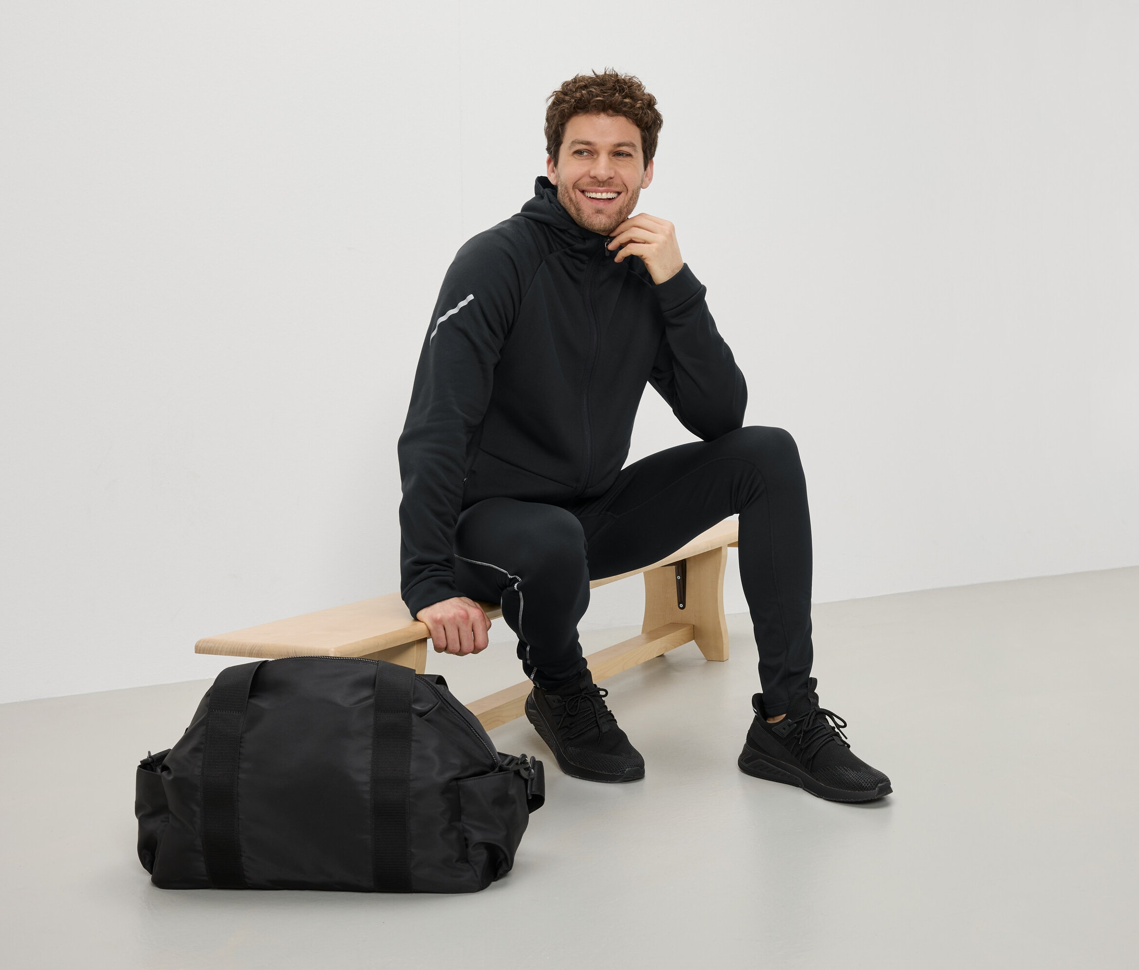 Férfi ül egy padon fekete sportruházatban, egy fekete sporttáska mellett.