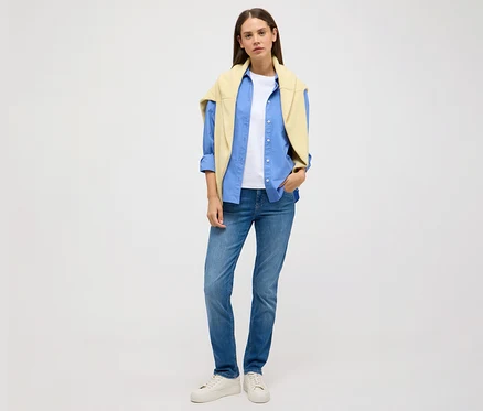 Nő áll világos háttér előtt, Angels »Cici Push-Up« slim fit farmernadrágot, kék inget, fehér pólót és sárga pulóvert visel.