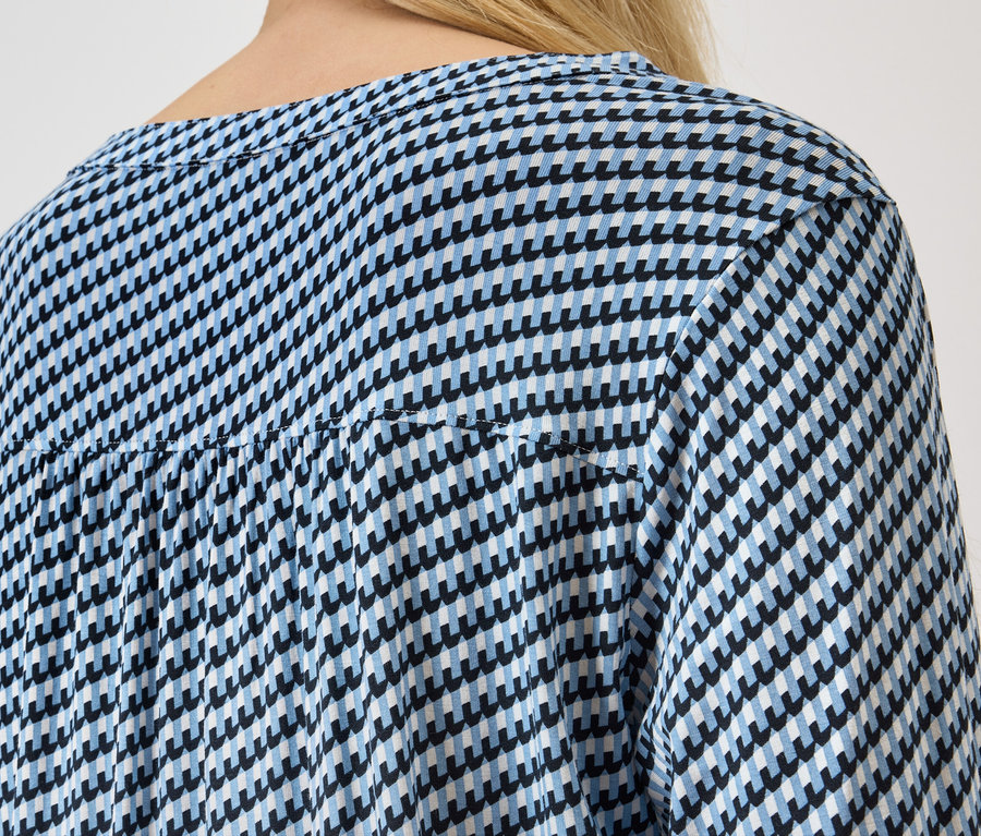 Hátulnézet egy szőke hajú nőről, aki kék-fehér, tyúkláb mintás blúzt visel.