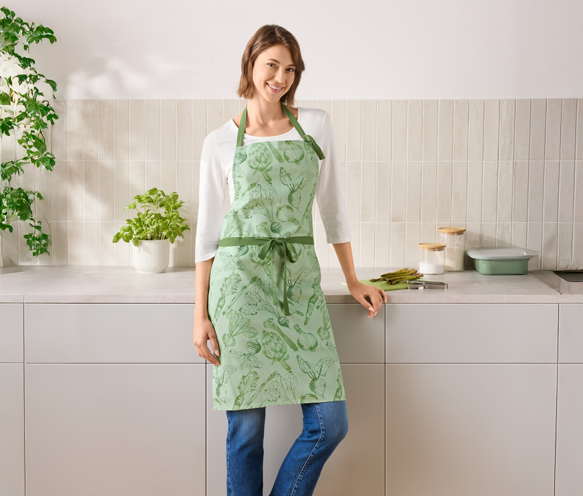 Egy nő áll a konyhában, zöld, zöldséges kötényt visel.