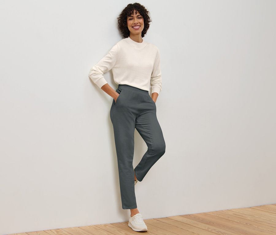 Egy nő a falnak dőlve pózol, szürke szabadidőnadrágot és pulóvert visel.