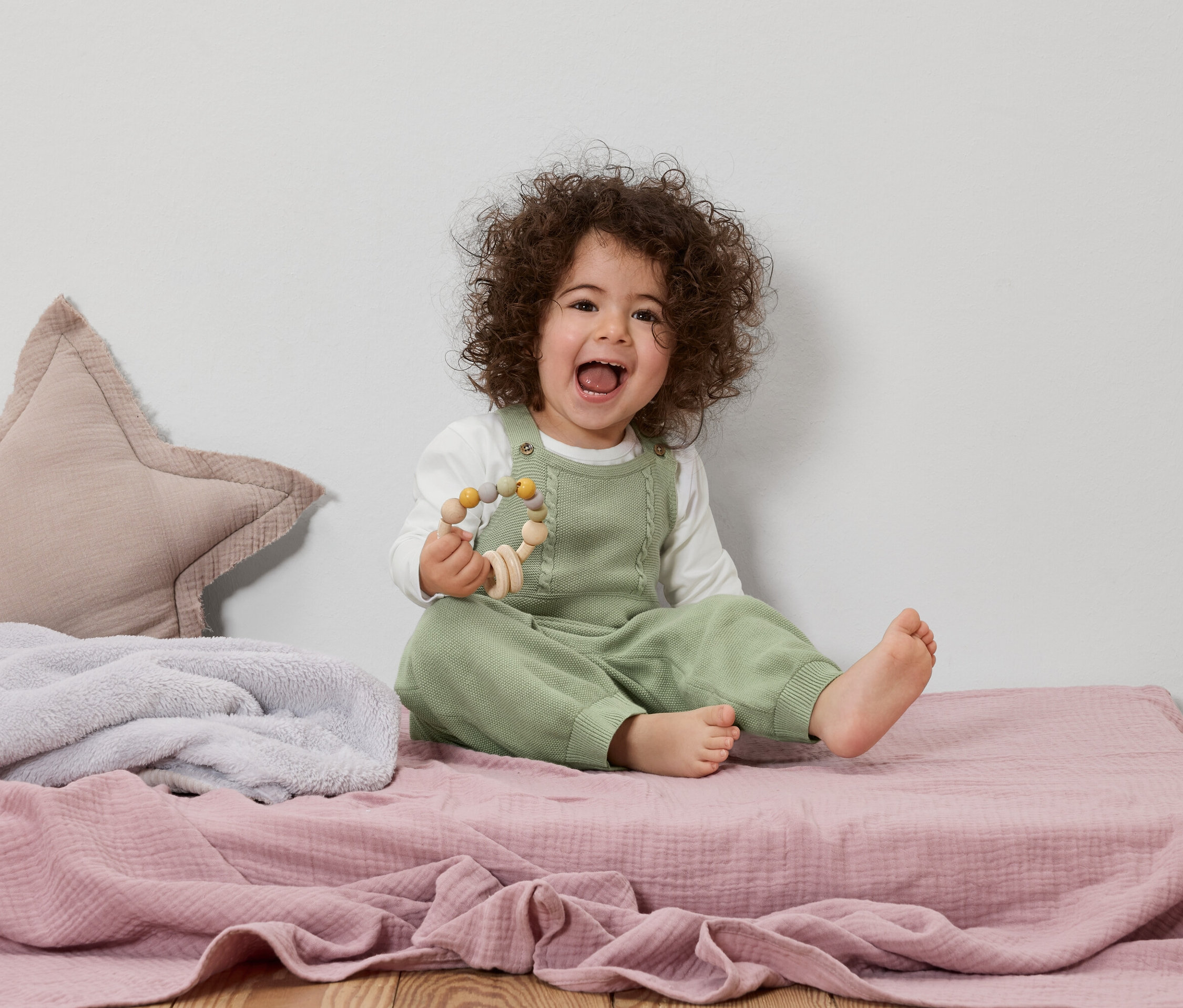 Egy göndör hajú, mosolygó kisgyermek ül egy rózsaszín játszószőnyegen, és egy fa játékot tart a kezében.