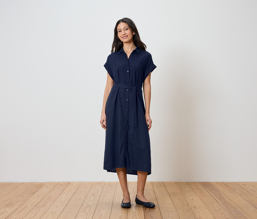 Egy nő áll a fal előtt, sötétkék ingruhát visel övvel és balerinacipővel.