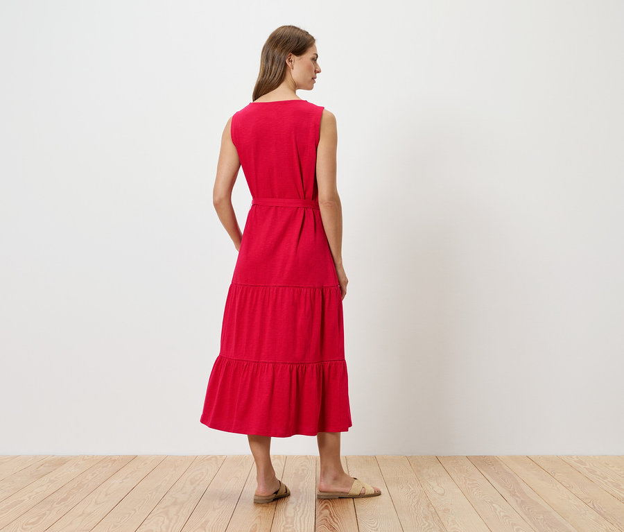 Hátulnézetből egy nő piros, ujjatlan, lépcsőzetes midi ruhában, övvel és lapos szandállal.
