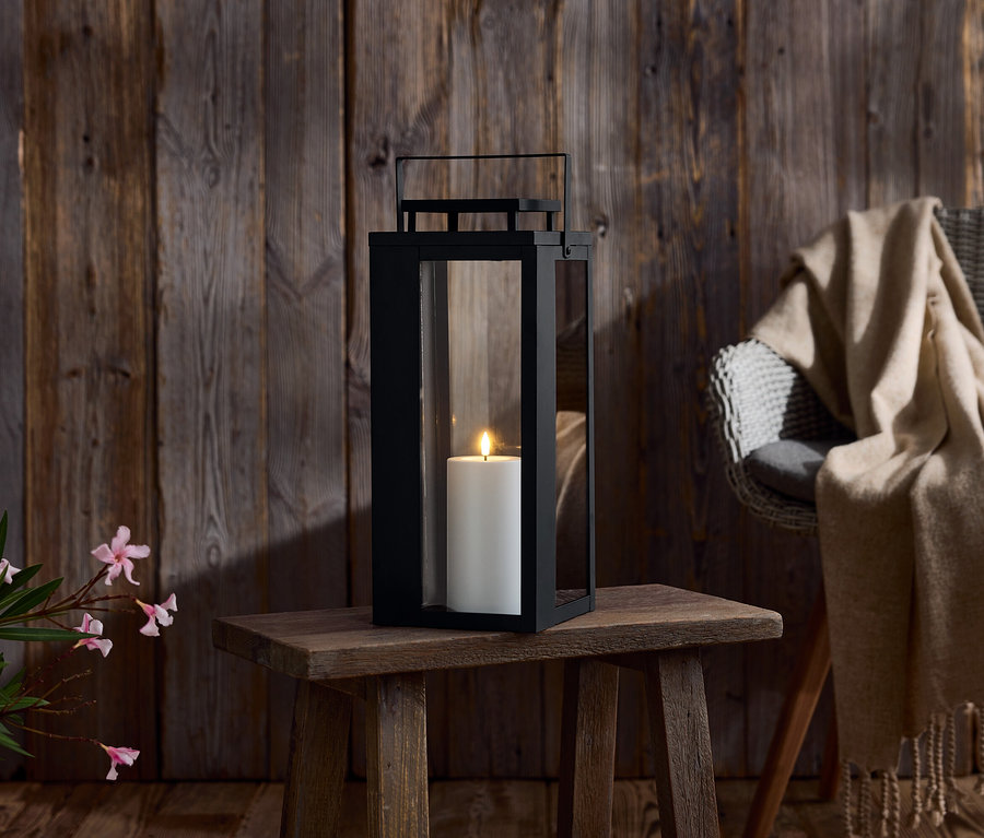 Egy fekete lámpás gyertyával egy fa asztalon. A háttérben fa fal és egy szék takaróval.